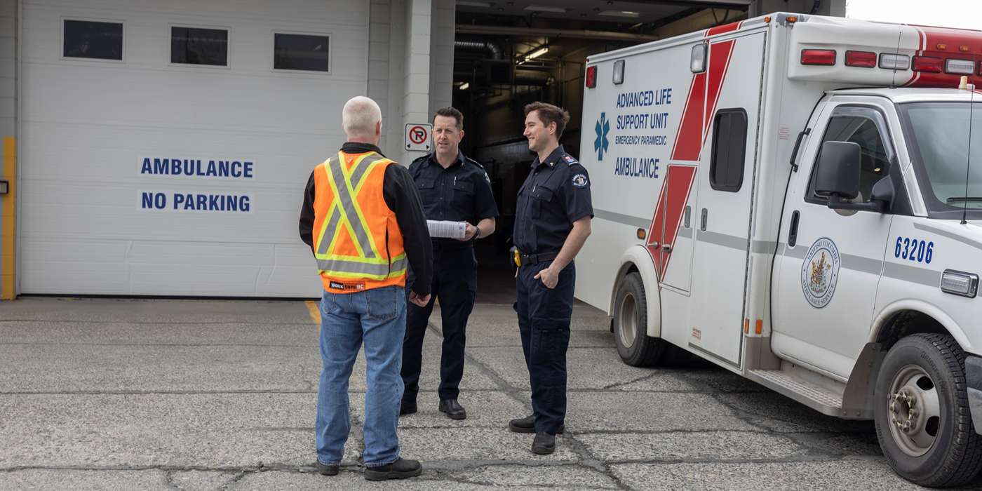 Standing next to an ambulance hall, two workers in dark blue uniforms and a worker in a high-visibility vest disuss work hazards