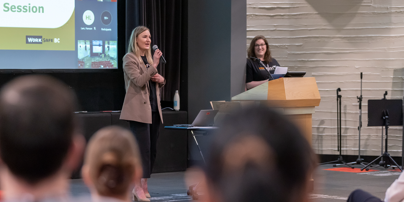 A person stands on stage holding a microphone and speaking to an audience during a WorkSafeBC information session. Another person stands behind a podium smiling, with a large presentation screen in the background displaying the WorkSafeBC logo.