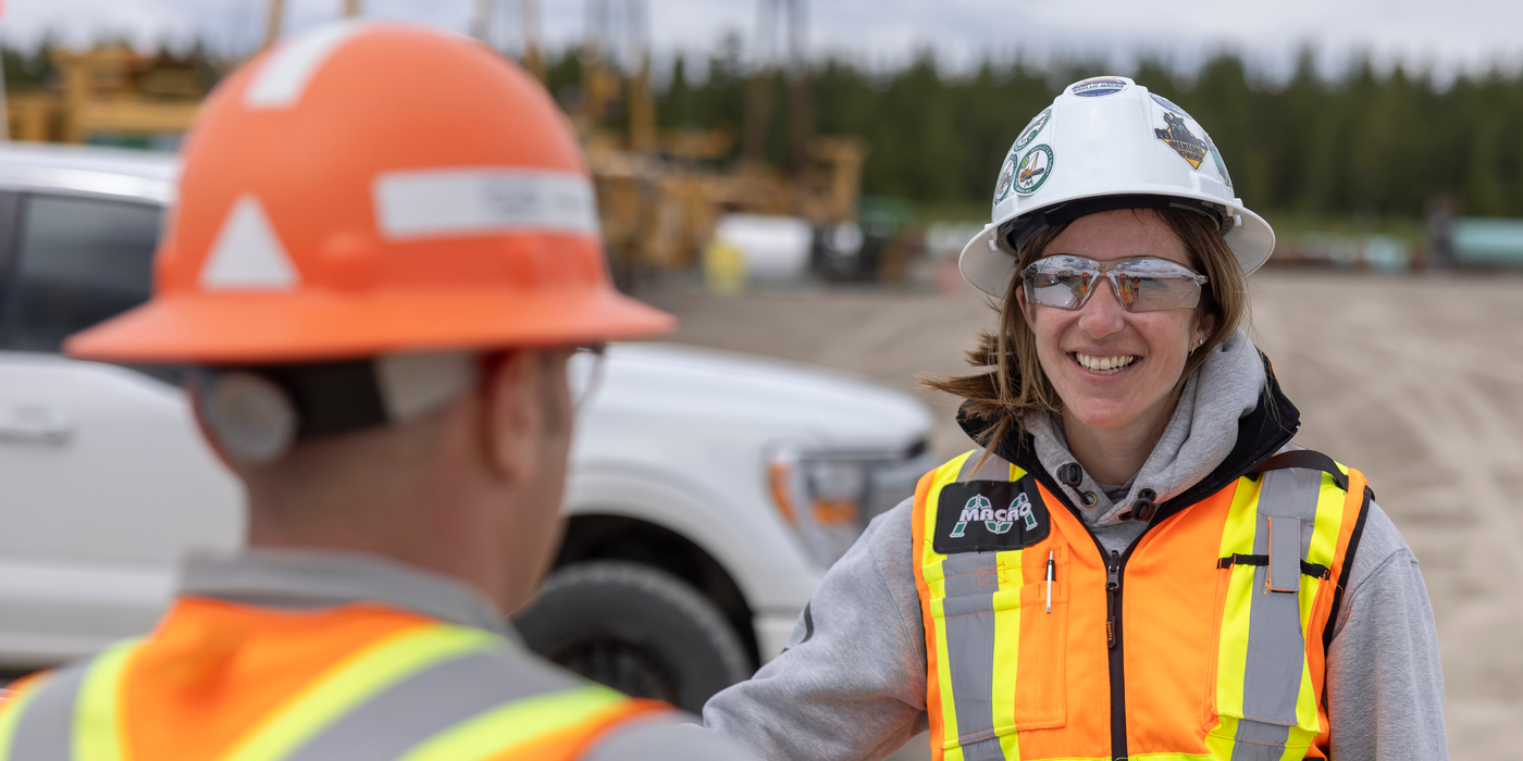 Two workers wearing high-visibility safety vests, hard hats, and safety glasses stand at an industrial worksite. One worker is in the foreground with their back to the camera, while the other faces the camera. Construction equipment, a pickup truck, and a forested background are visible behind them.