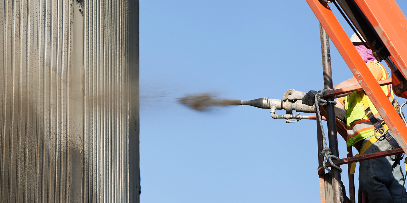 Worker wearing high‑visibility safety gear standing on an elevated platform, operating a spray nozzle to apply shotcrete onto a vertical concrete surface, with a clear blue sky in the background.