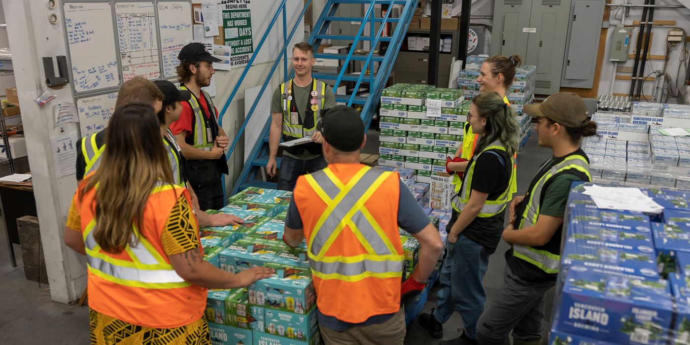 Group of workers in high-visibility yellow and orange vests, meeting in a warehouse with stacks of goods around them