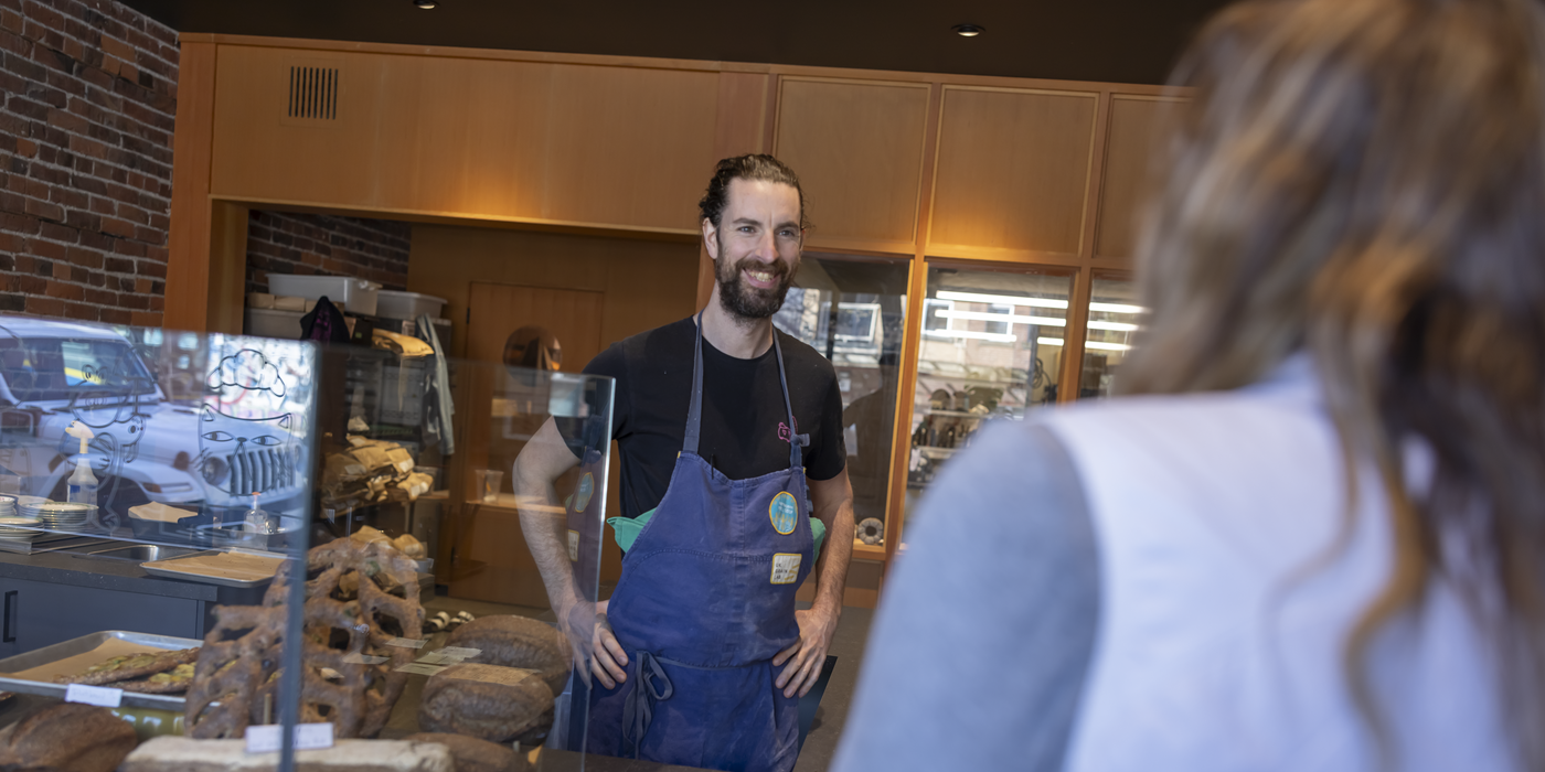Photo of smiling man in apron behind bakery counter, serving customer