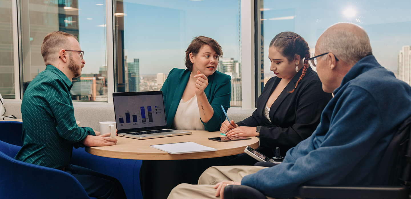 Four workers of different ages, dressed in casual business style, are at a conference table, using tools like a laptop, a tablet, a pad of paper, and an assistive device. 